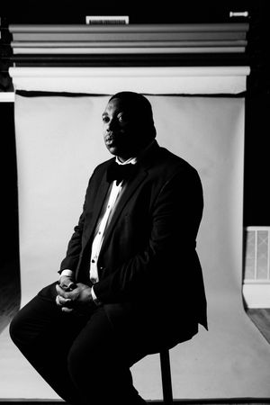 Black and white portrait of a man in a tuxedo sitting gracefully in a studio setting.