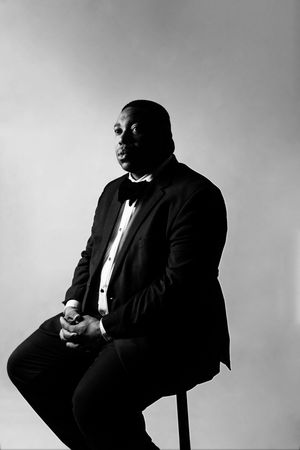 Black and white portrait of a man in a tuxedo with bow tie seated against a light background.
