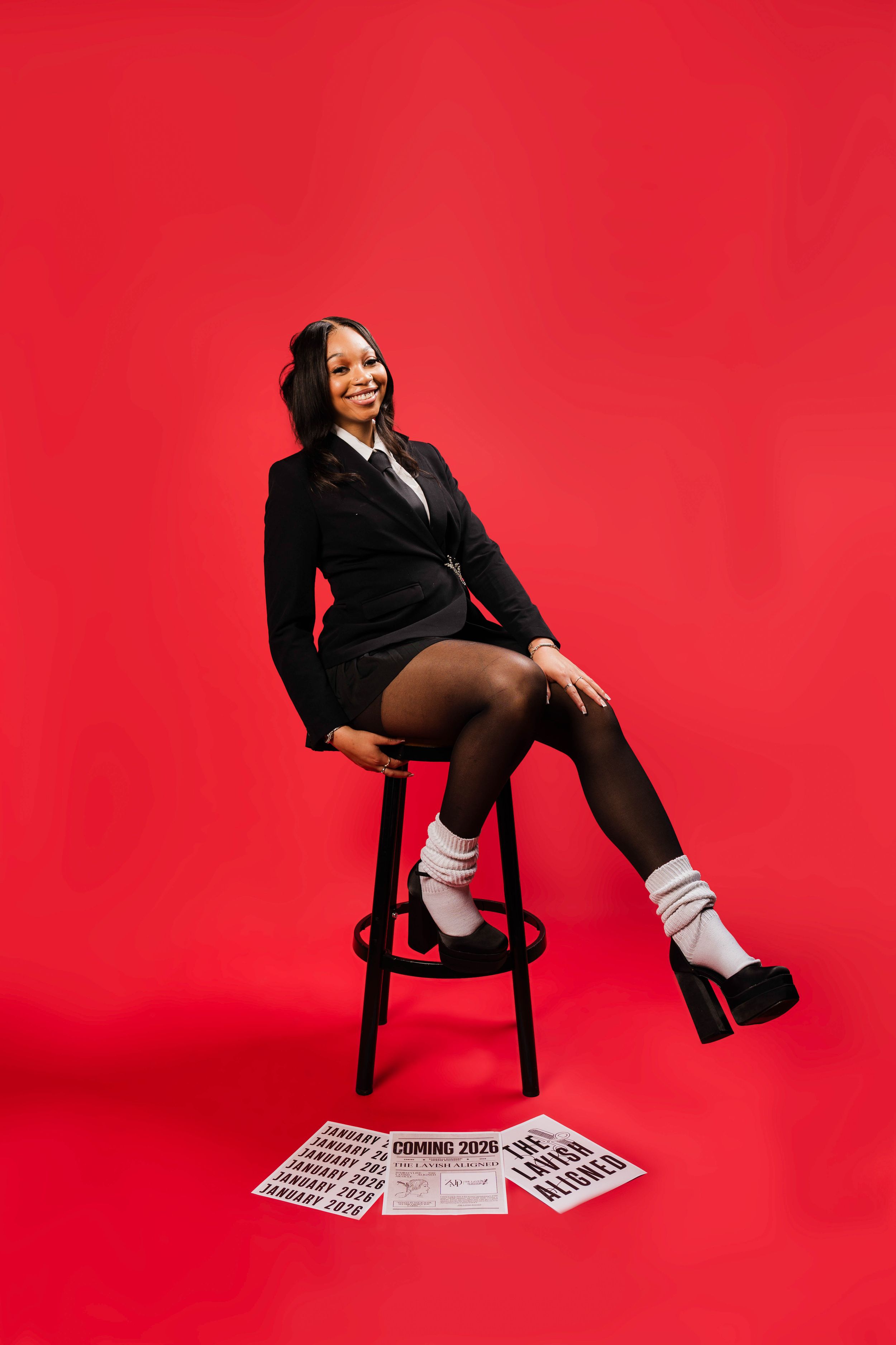Smiling woman in black suit sits on stool against red background with newspapers on floor.