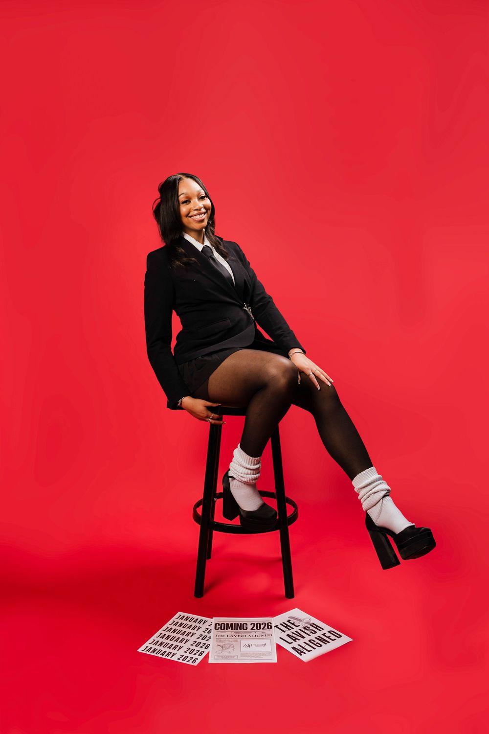 Smiling woman in black suit sits on stool against red background with newspapers on floor.