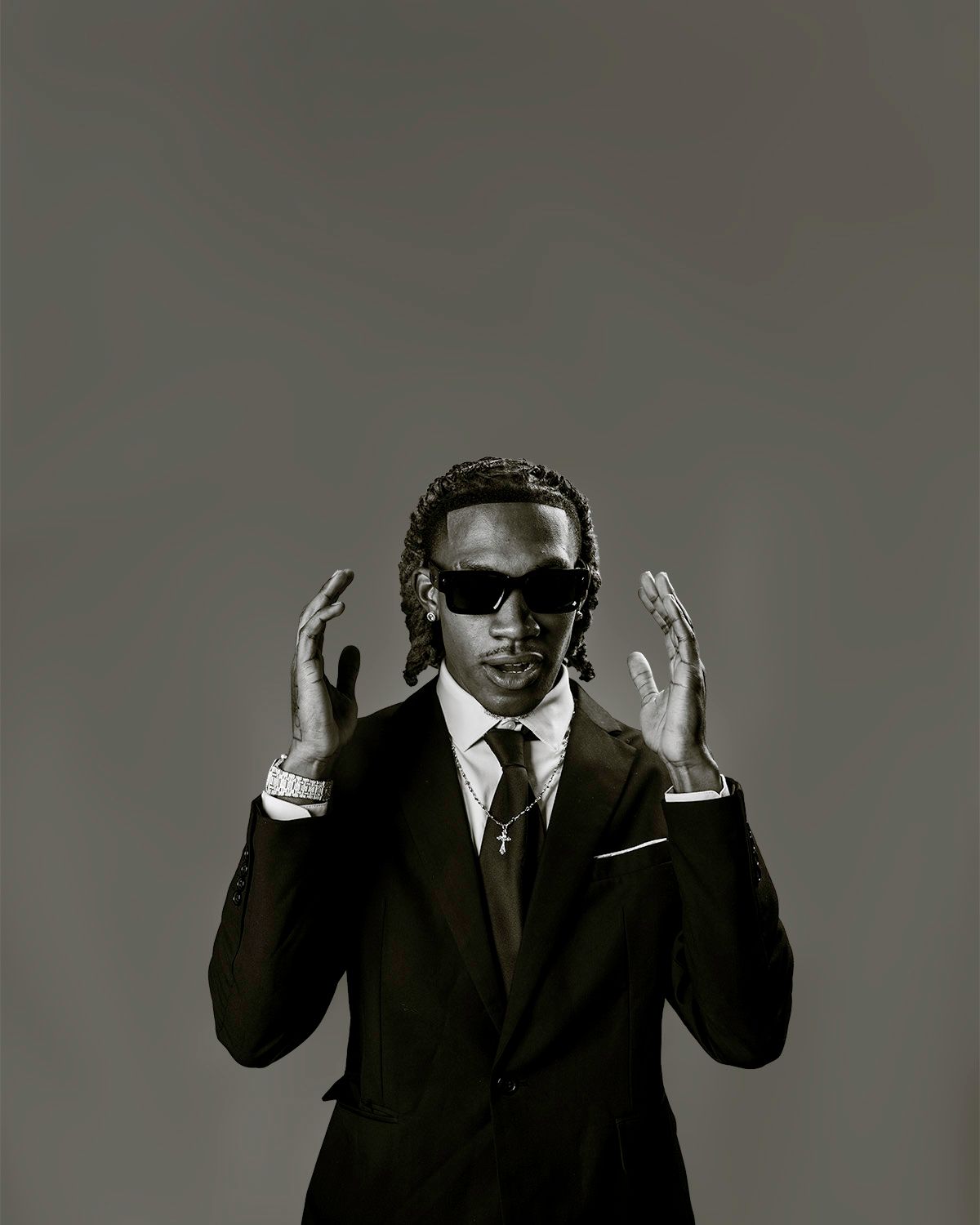 Man in black suit and sunglasses with braids poses confidently in a black and white portrait photo.