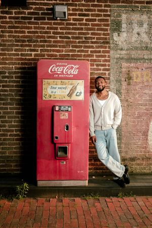Man leaning against a vintage red Coca-Cola vending machine on a brick wall backdrop.