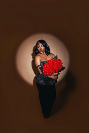 Woman in black lace dress holding large red rose bouquet under spotlight on brown background.
