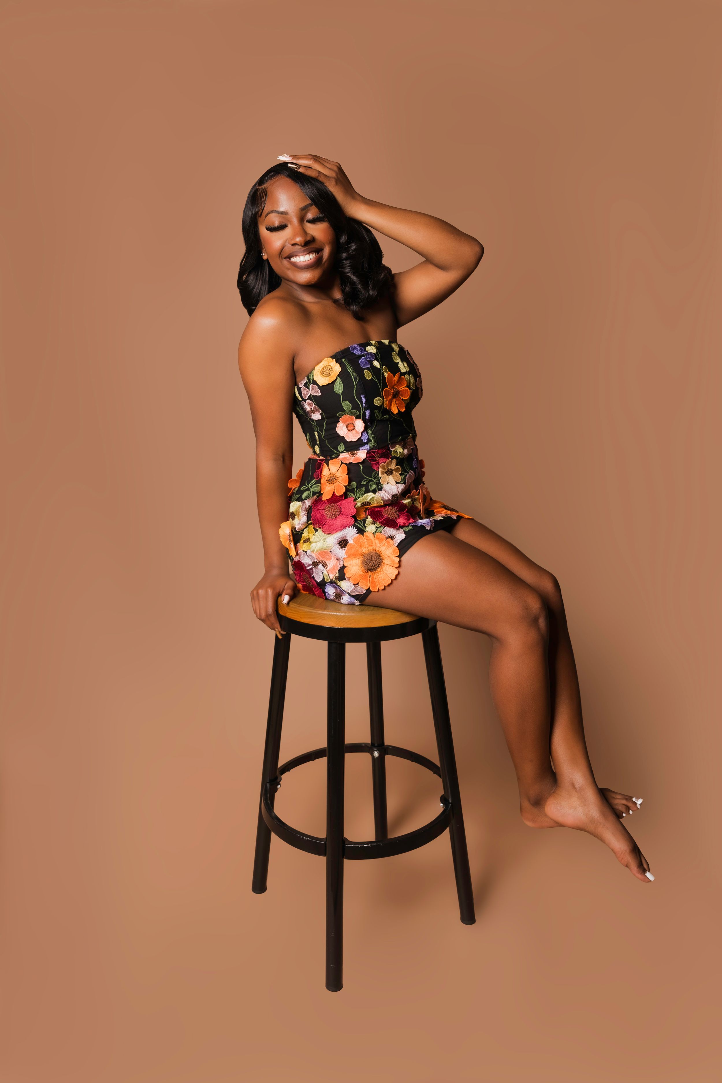 Smiling Black woman in floral strapless mini dress sits on wooden bar stool against warm brown background.