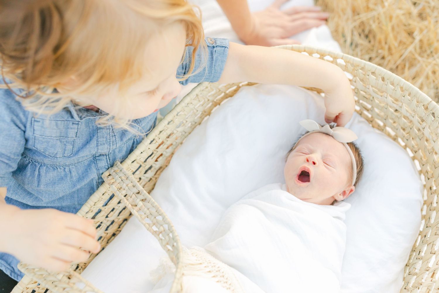 Newborn baby girl swaddled in white blanket with bow headband lying in a wicker Moses basket.