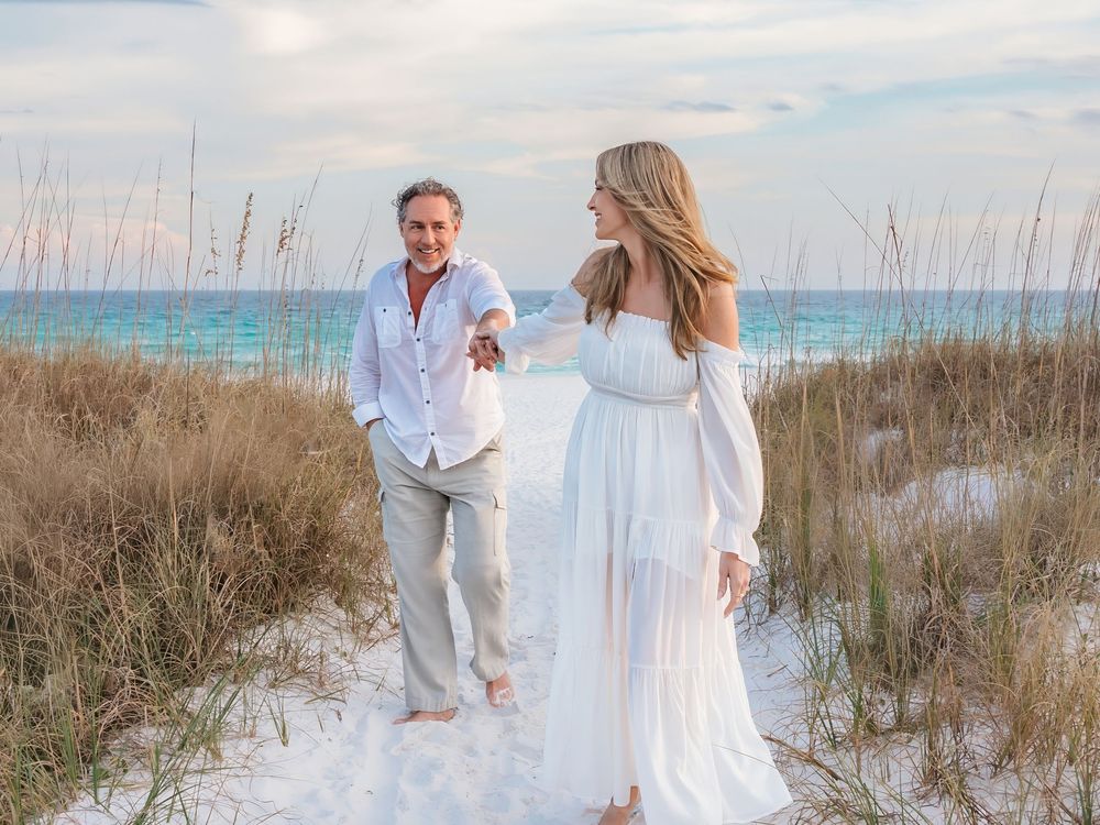 Romantic couple walking through beach dunes at sunset, woman in flowing white off-shoulder dress.