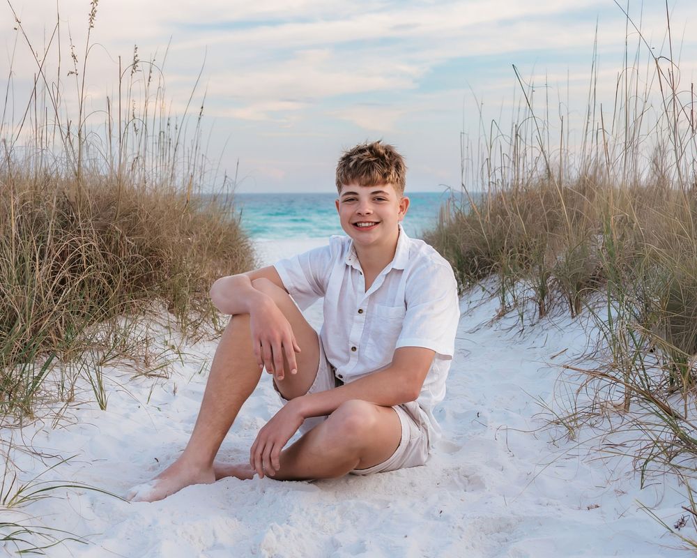 Smiling teenage boy sitting in white sand dunes surrounded by sea grass at the beach.