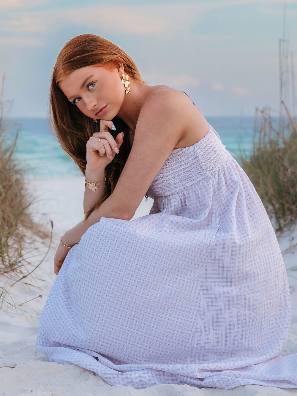 Young woman in lavender gingham dress posing on sand dunes with ocean in the background.