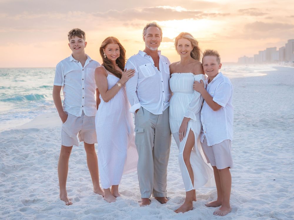 Family of five in coordinating white and tan outfits posing on beach during vibrant sunset.