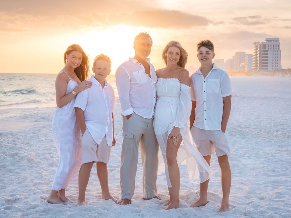 Family of five smiling together on sandy beach during vibrant orange sunset, wearing white outfits.