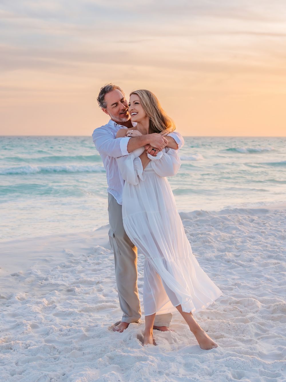 Couple dancing joyfully on white sand beach at sunset, woman in flowing white dress laughing.