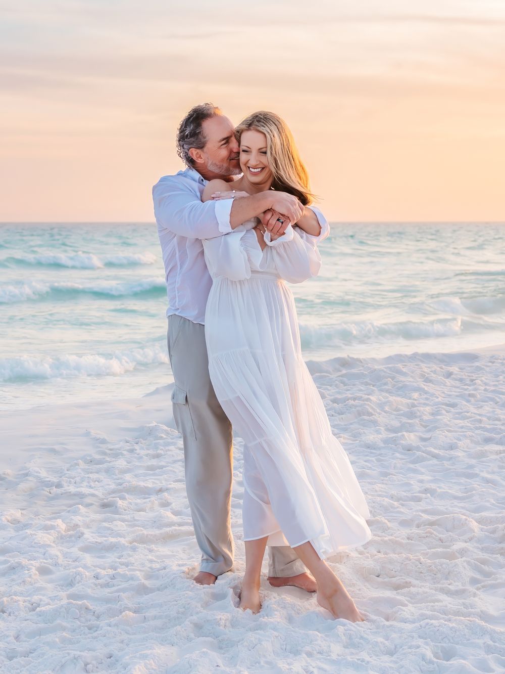 Couple embracing on white sand beach at sunset, woman in flowing white dress, man in light blue shirt.