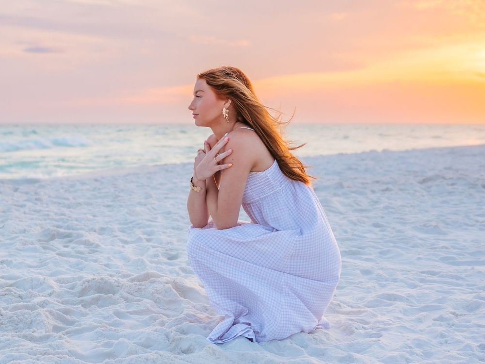 Young woman in white dress crouching on beach at pastel sunset, gazing thoughtfully at the ocean.