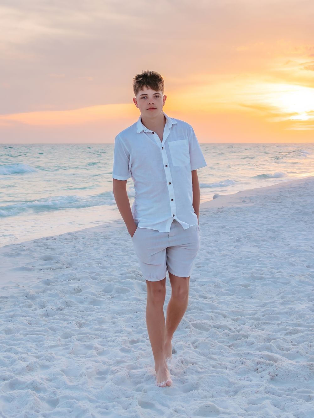 Teen boy walking barefoot on beach at golden sunset, wearing white button-up shirt and gray shorts.