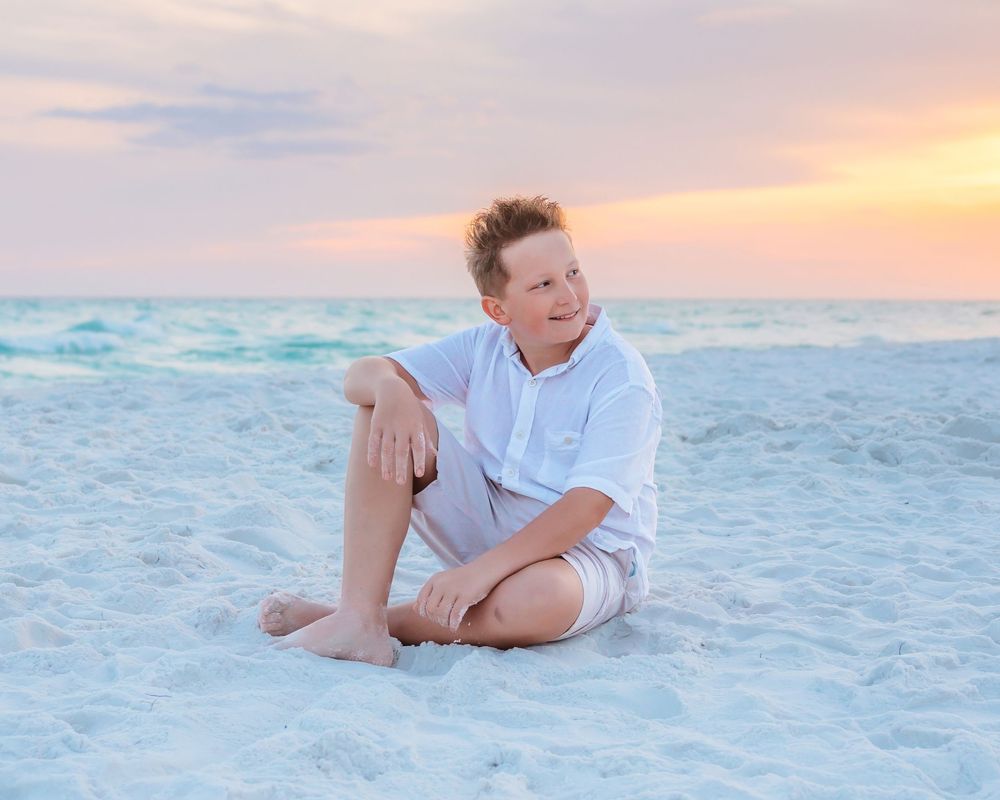 Young boy sitting on white sand beach at colorful sunset, wearing white shirt and khaki shorts.