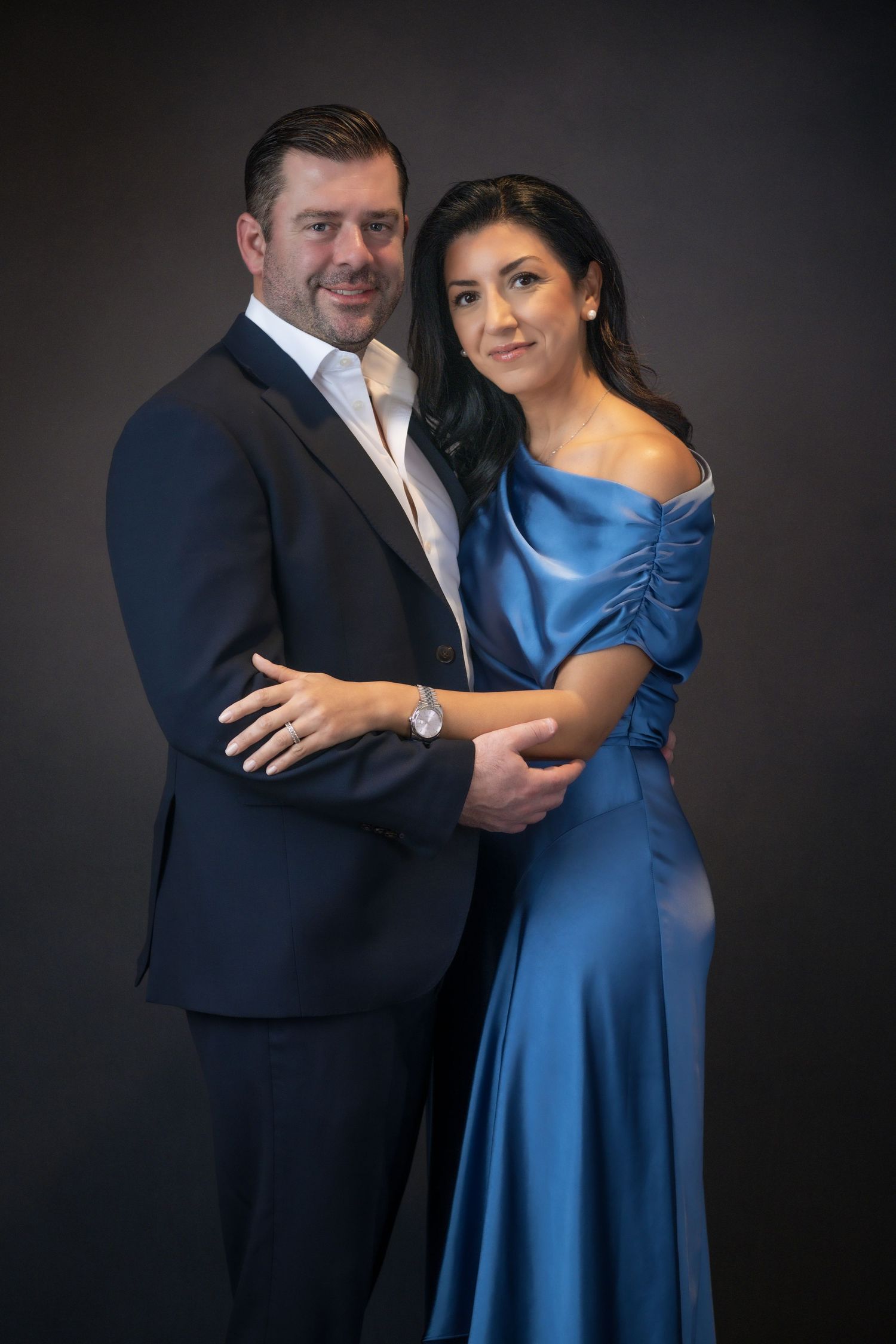 Elegant couple posing in formal attire, man in dark suit, woman in blue satin off-shoulder gown, studio portrait.