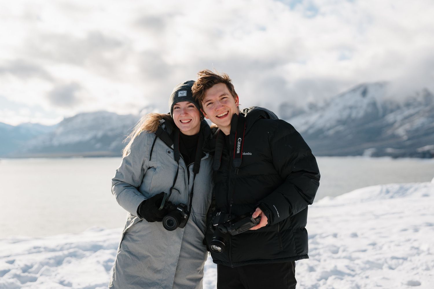 A winter scene with two people in warm coats standing together against a snowy mountain backdrop.