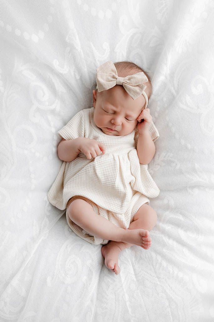 Sleeping newborn baby girl in cream knit outfit and bow headband lying on white textured blanket.