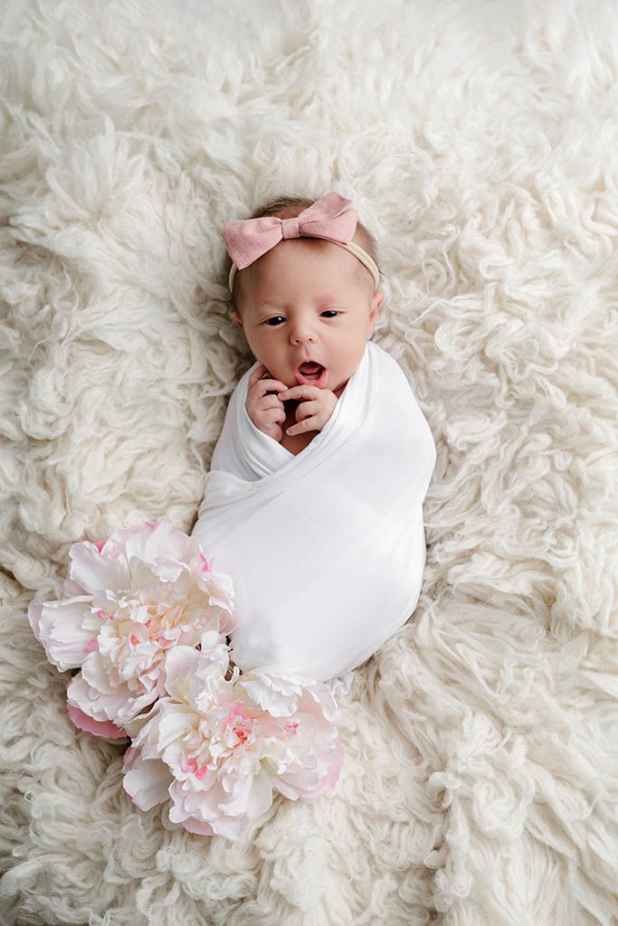 Newborn baby girl swaddled in white wrap with pink bow headband and peony flower on fluffy cream rug.