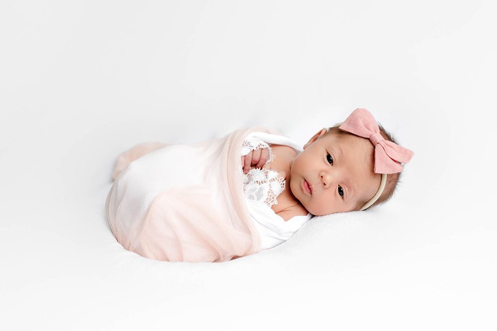 Newborn baby girl swaddled in pink blanket with bow headband lying on white background.