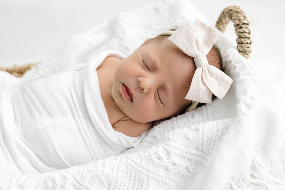 Sleeping newborn baby wrapped in white swaddle with bow headband in a cozy basket.