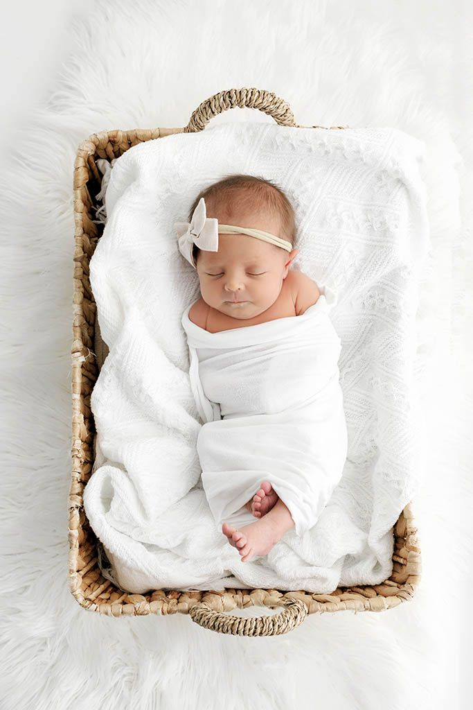 Sleeping newborn baby girl swaddled in white wrap with bow headband lying in wicker basket on fluffy white fur.