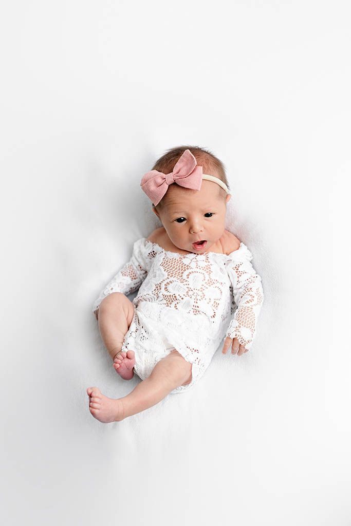 Newborn baby girl in white lace romper and pink bow headband lying on white background.