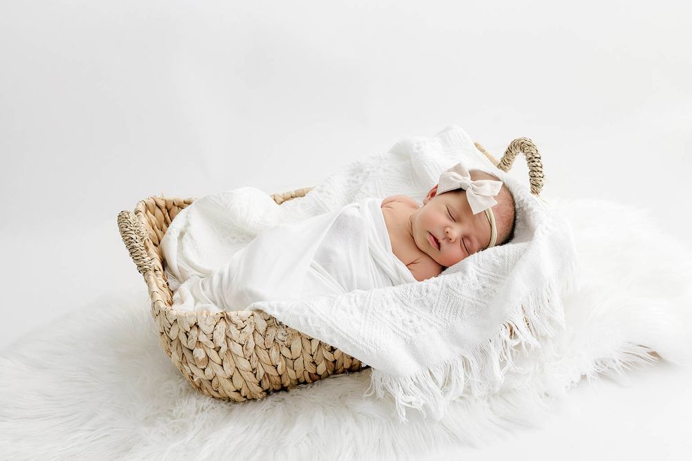 Sleeping newborn baby wrapped in white swaddle with bow headband resting in a wicker basket on fluffy white fur.