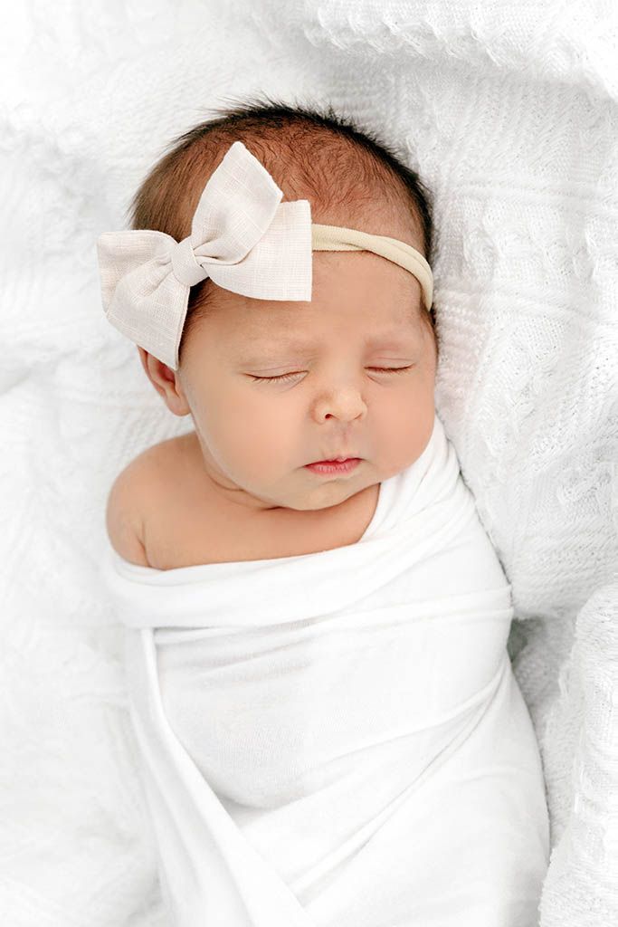Close-up of peaceful sleeping newborn baby girl swaddled in white wrap wearing a cream bow headband.
