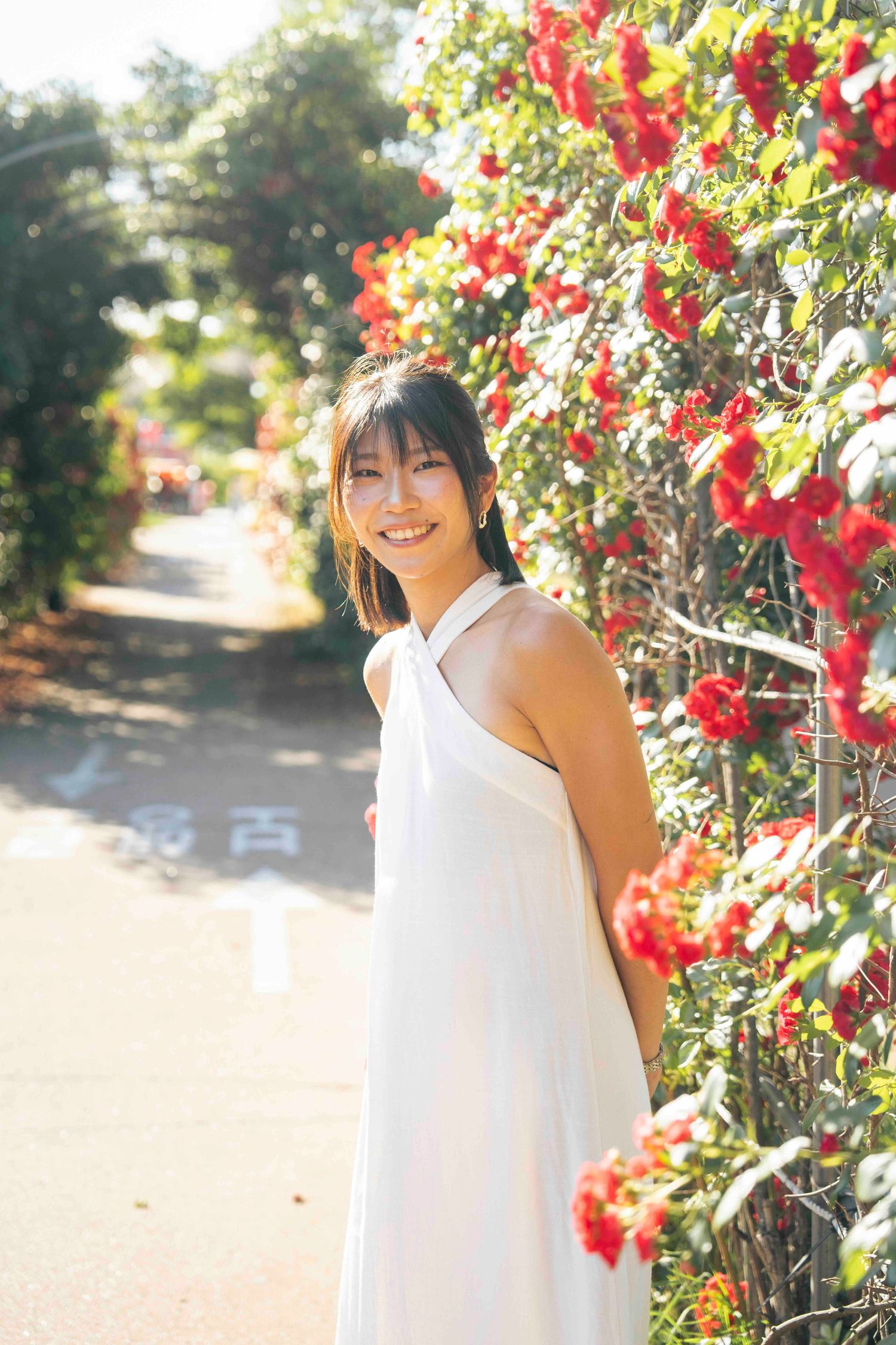 A smiling young woman in a white halter dress stands beside vibrant red flowering bushes on a sunlit garden path.