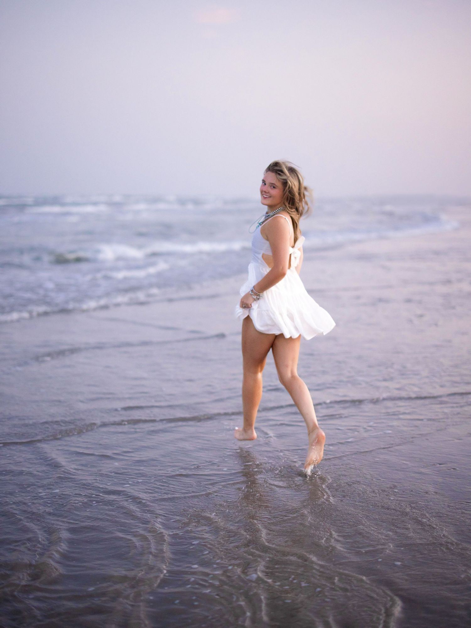 Woman in white dress walking barefoot on a sandy beach with ocean waves in the background at dusk.