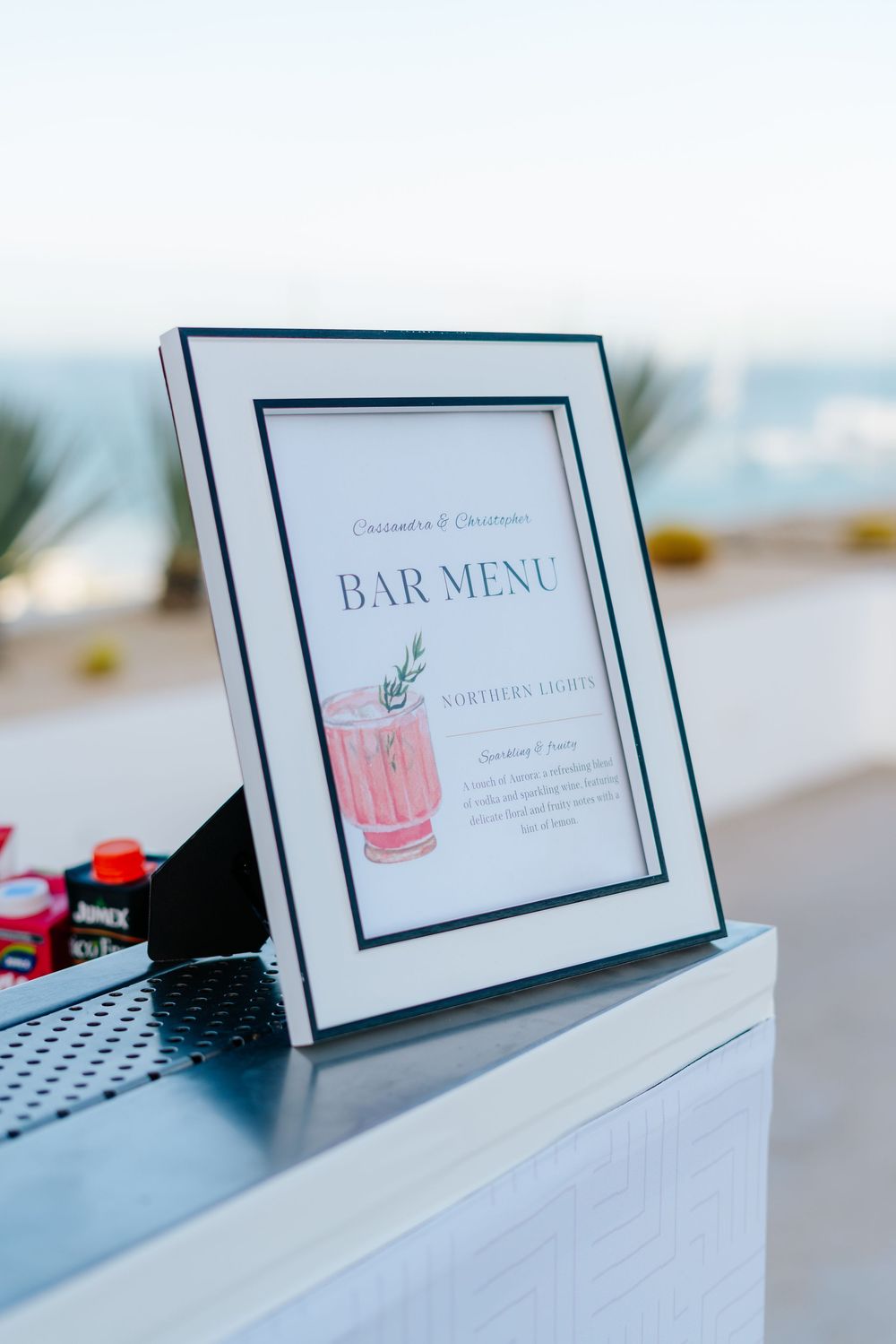 Elegant framed bar menu sign displayed on a white counter at an outdoor wedding reception overlooking the ocean.