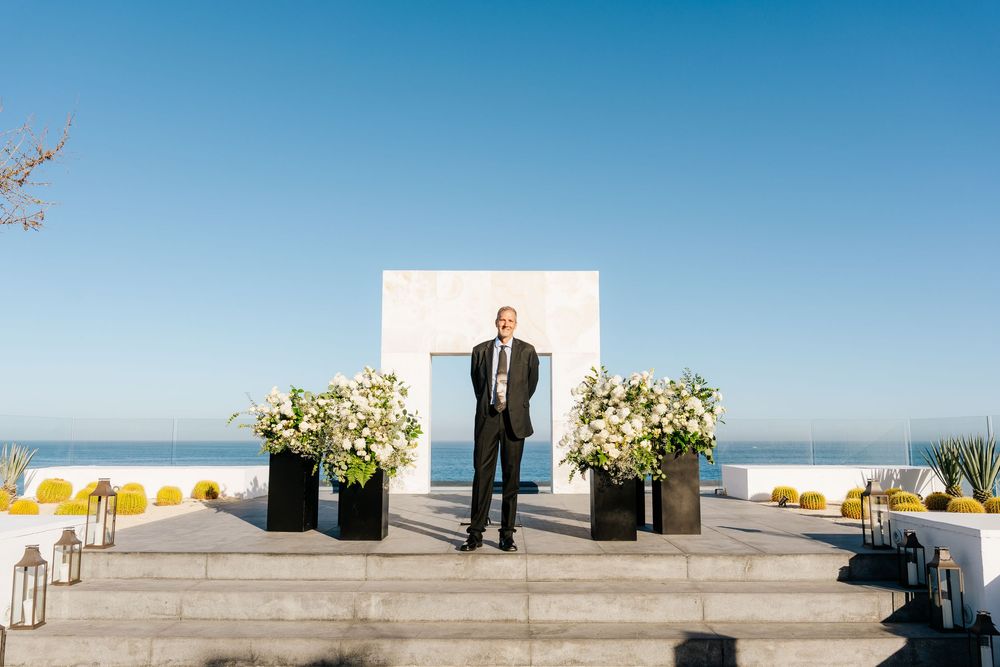 Man in suit stands at elegant outdoor wedding altar with white floral arrangements and ocean view backdrop.