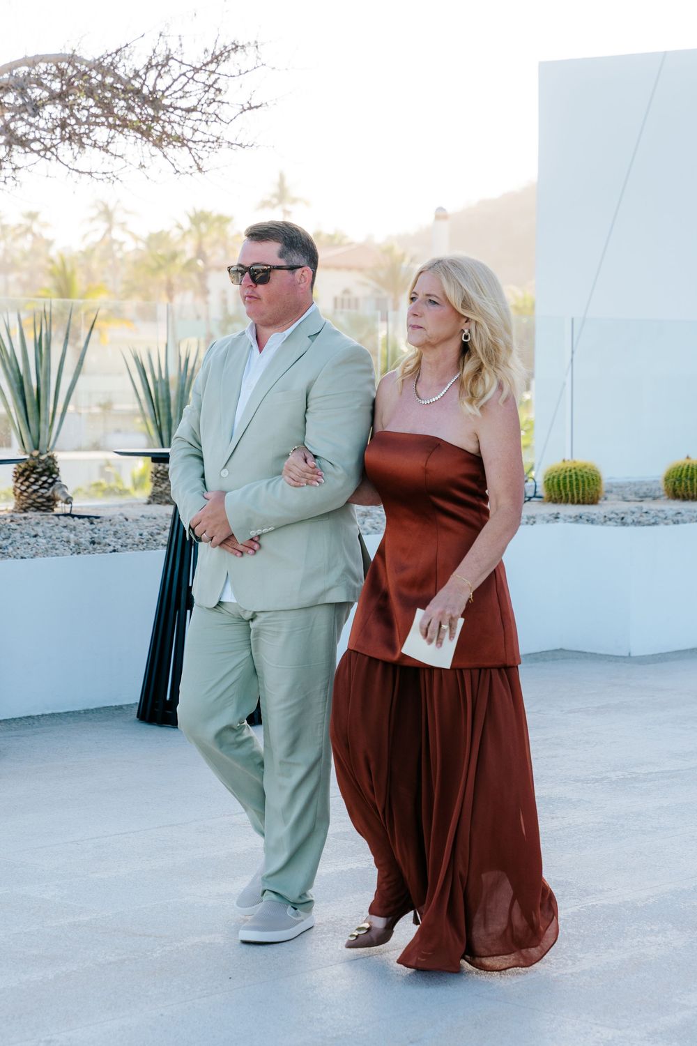 Elegant couple walking outdoors, man in sage suit, woman in rust strapless gown, desert landscape background.