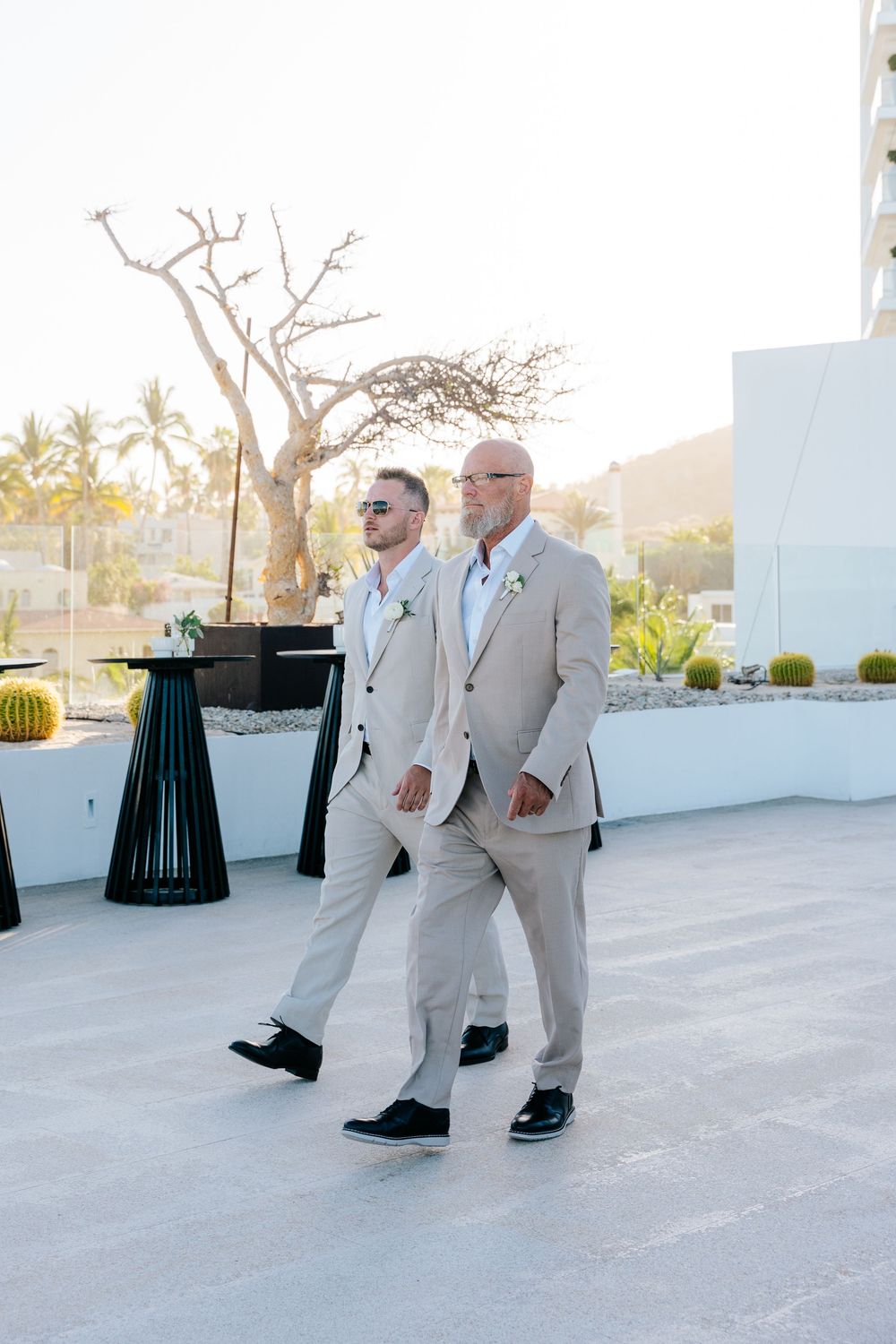 Two men in matching tan suits walk together at an outdoor wedding ceremony with tropical scenery.