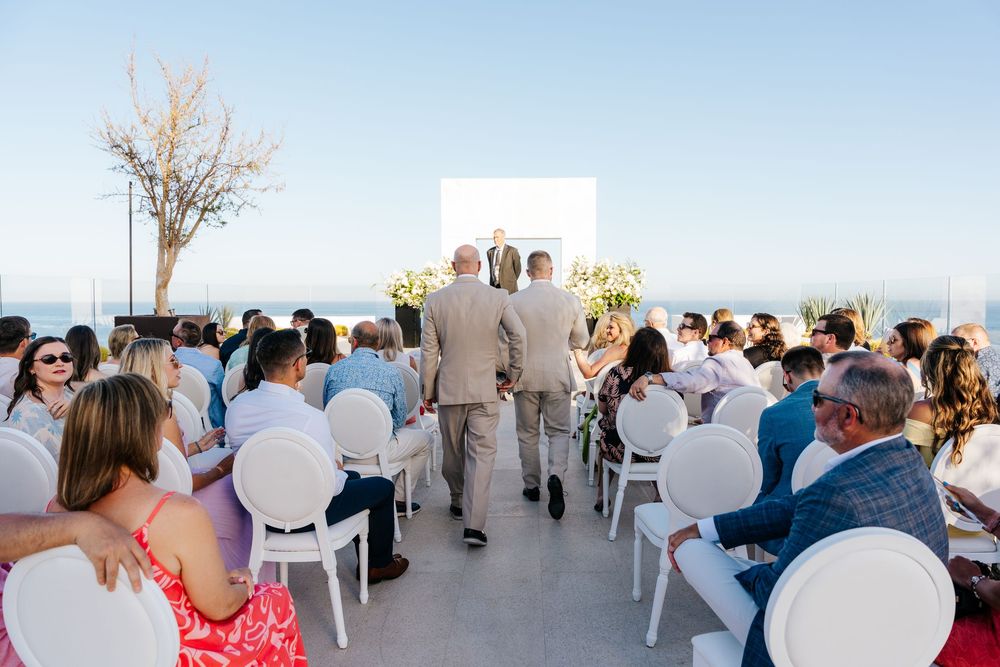 Outdoor wedding ceremony with guests seated in white chairs overlooking the ocean on a sunny day.
