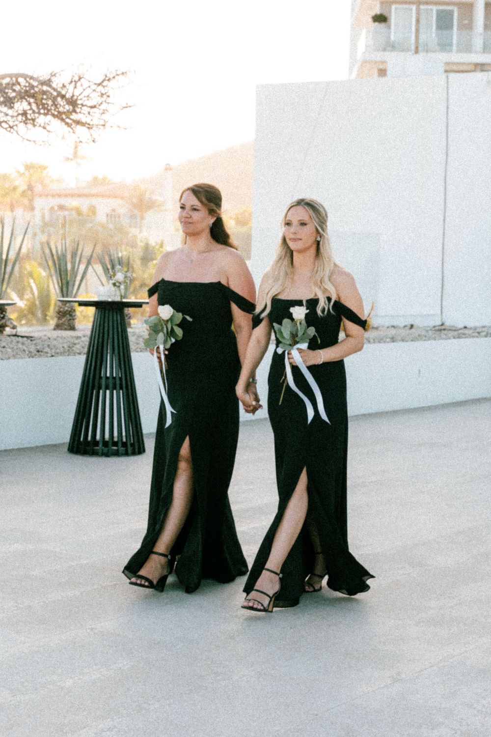 Two bridesmaids in black off-shoulder gowns with slits walk together holding flowers at an outdoor wedding.
