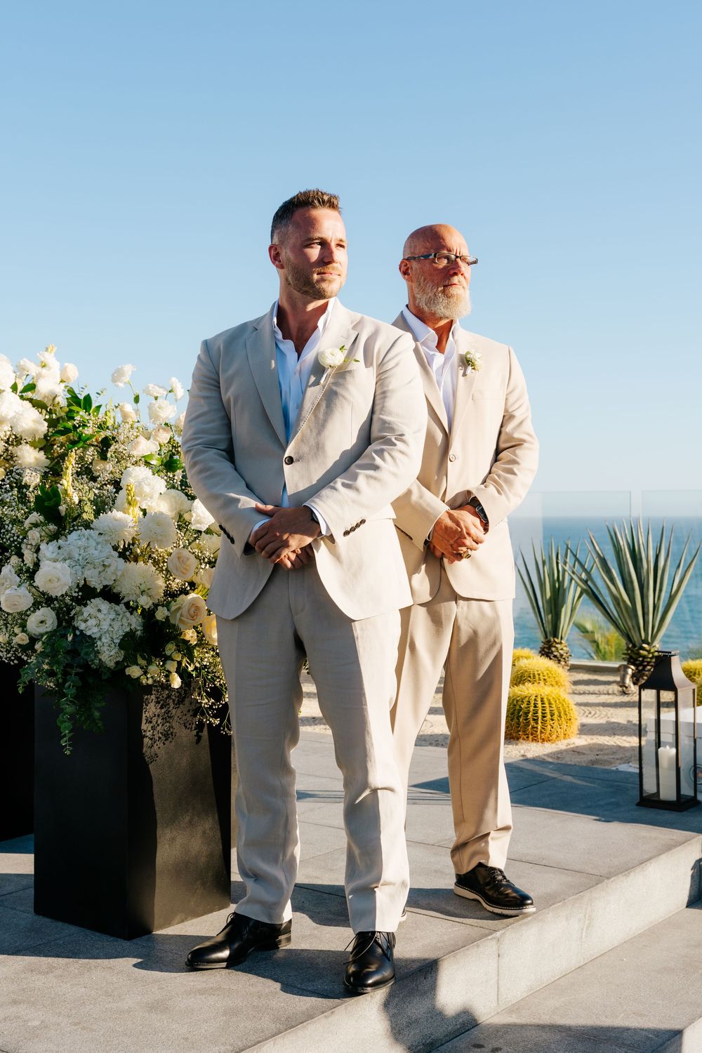 Two grooms in matching beige suits stand at the altar with white floral arrangements and ocean backdrop.