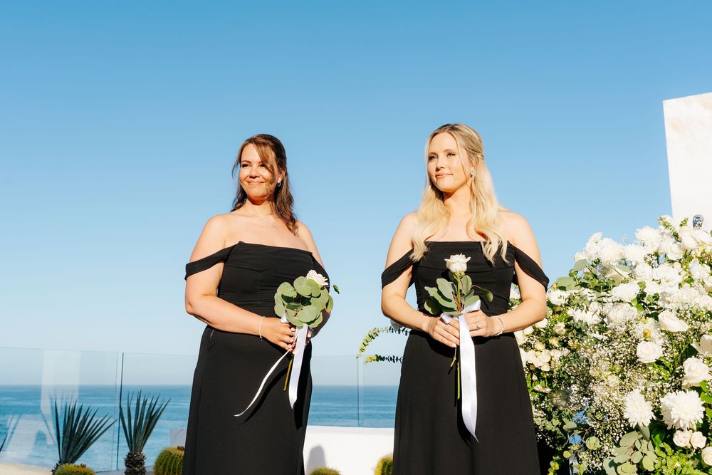 Two bridesmaids in black off-shoulder gowns holding white roses at an outdoor wedding ceremony.