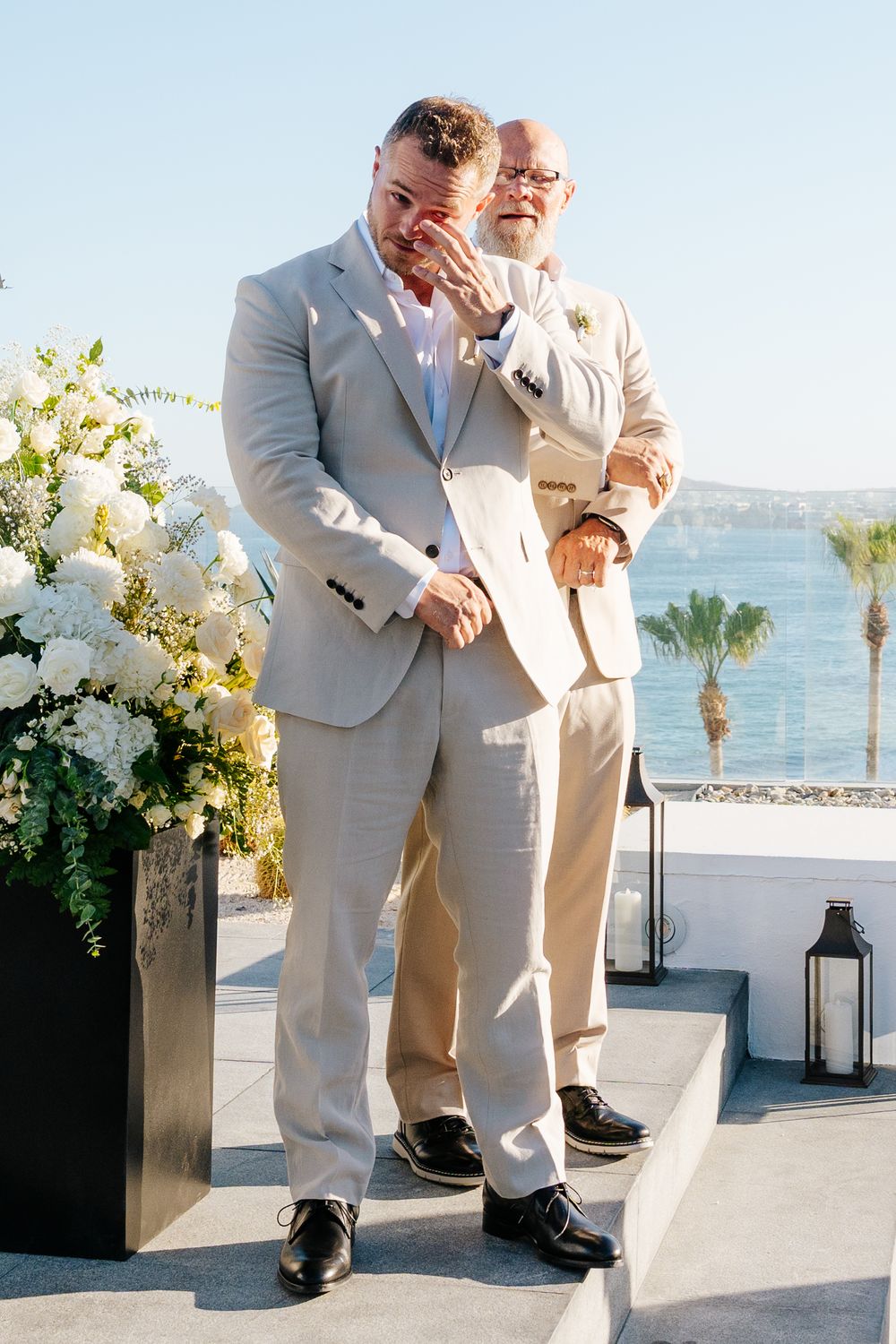Emotional groom wiping tears at outdoor wedding ceremony with white floral arrangements and ocean backdrop.
