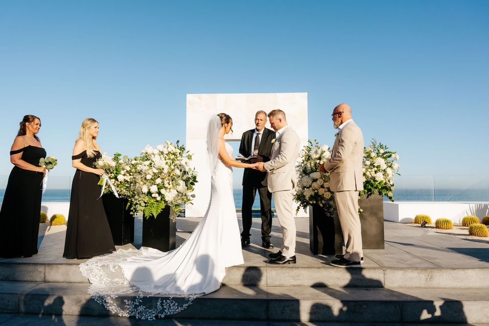 Bride and groom exchange vows at an outdoor wedding ceremony with white floral arch and ocean backdrop.