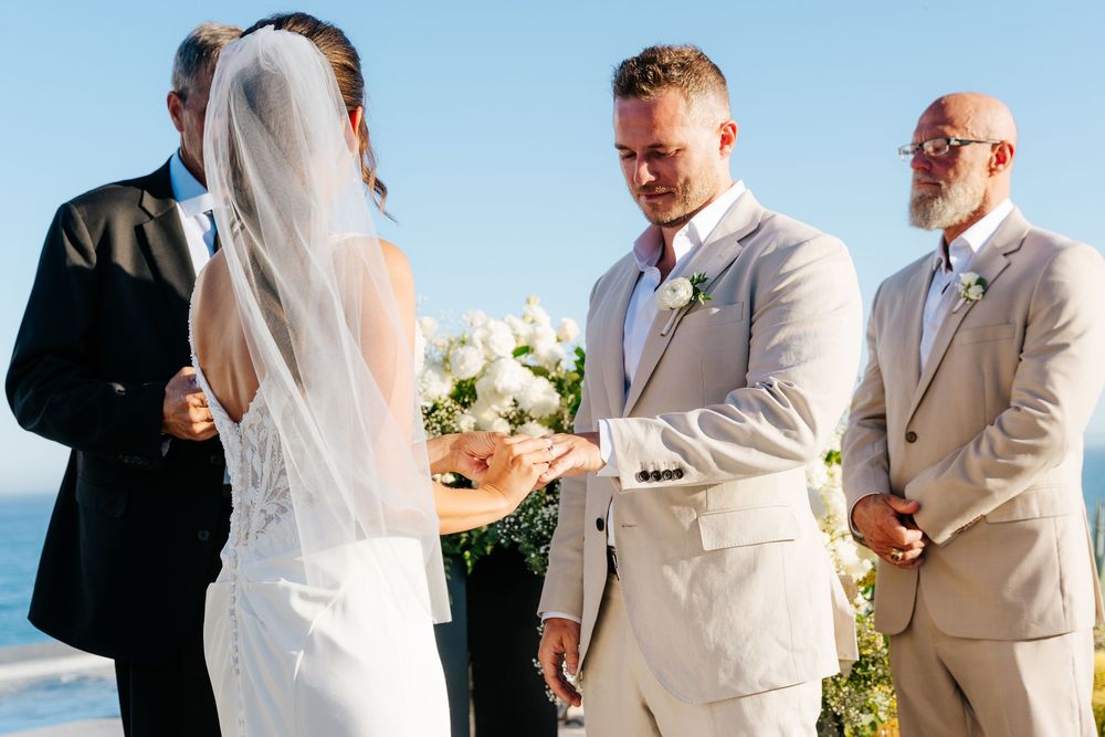 Groom in beige suit holds bride's hands during an outdoor wedding ceremony by the ocean.