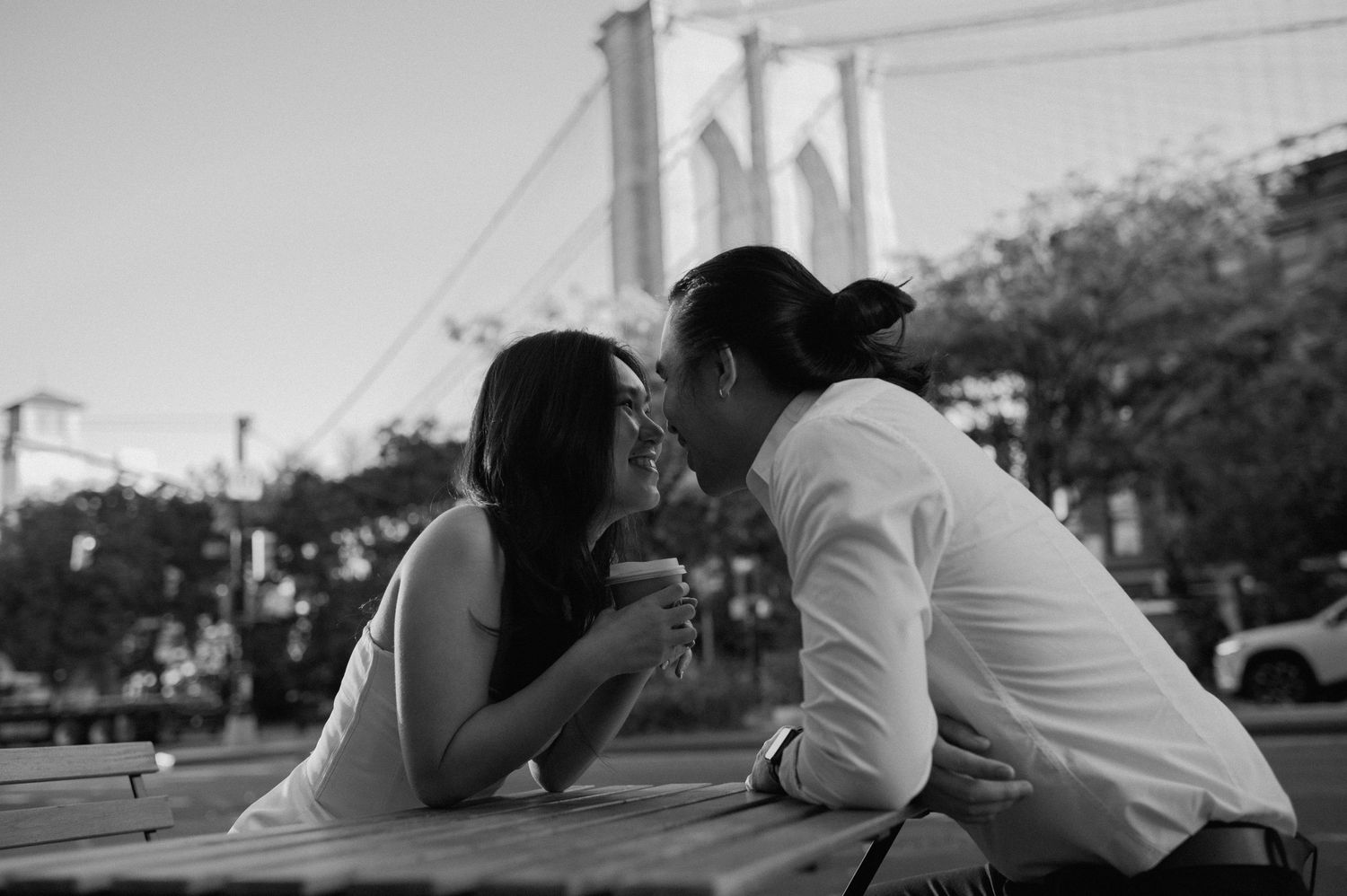 Couple sharing a romantic moment at an outdoor table with Brooklyn Bridge in the background, black and white photo.