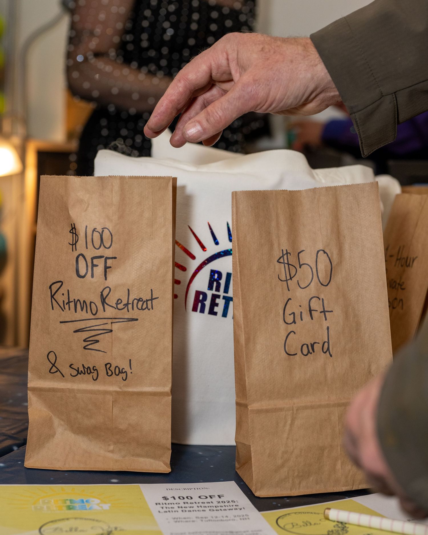 Brown paper bags labeled '$100 Off Ritmo Retreat & Swag Bag' and '$50 Gift Card' at a raffle table at latin dance social.