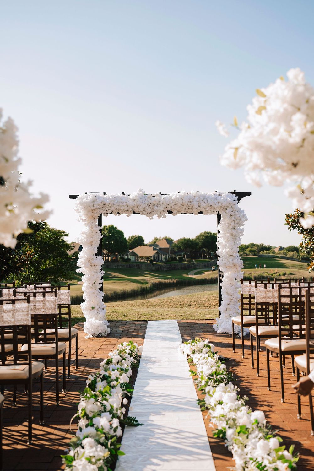 Elegant outdoor wedding ceremony setup with white floral arch, white aisle runner, and wooden chairs on a golf course.