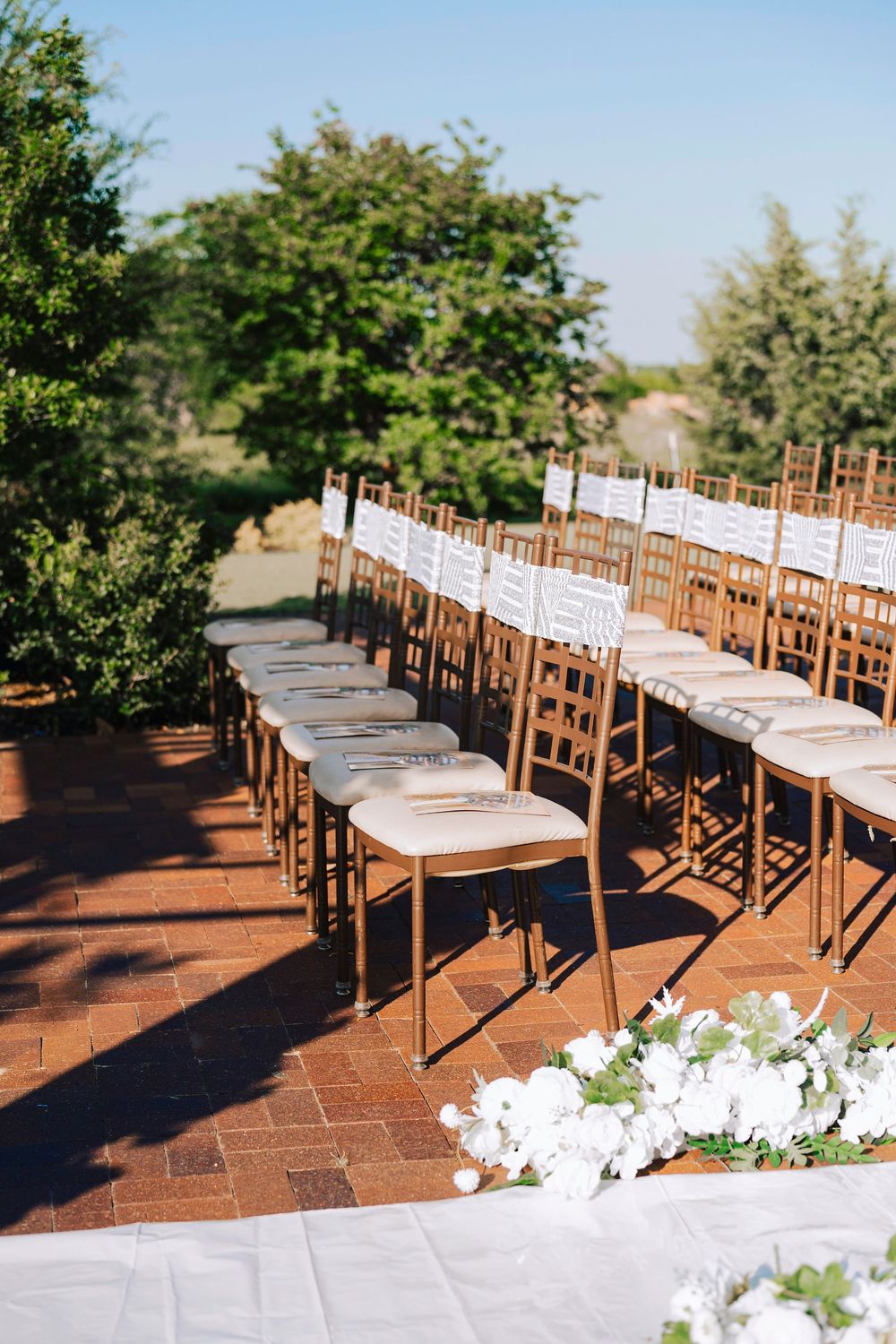 Rows of gold Chiavari chairs arranged on brick patio for outdoor wedding ceremony, adorned with white floral accents.