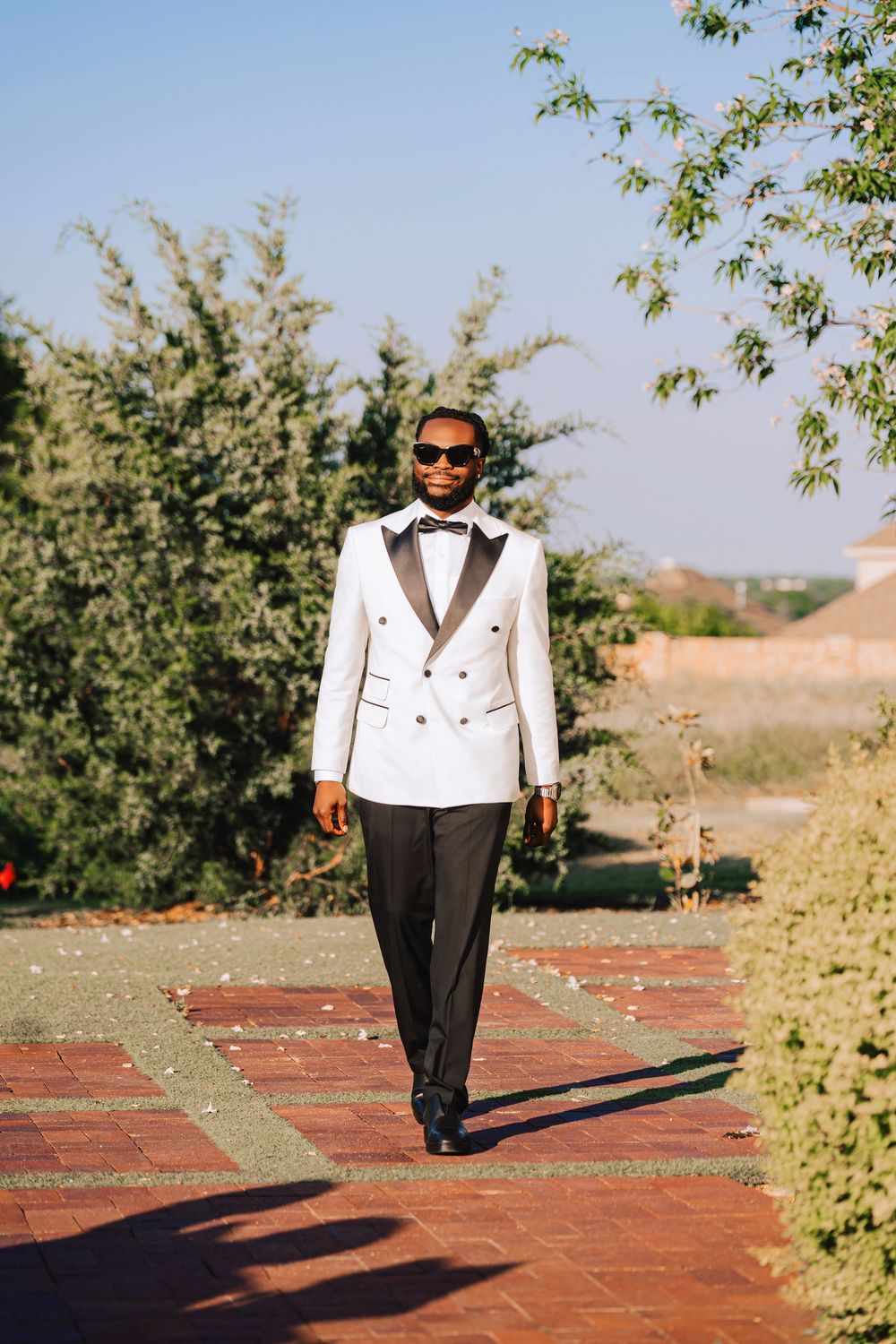 Stylish groom in white double-breasted tuxedo and sunglasses walks along a brick garden path.