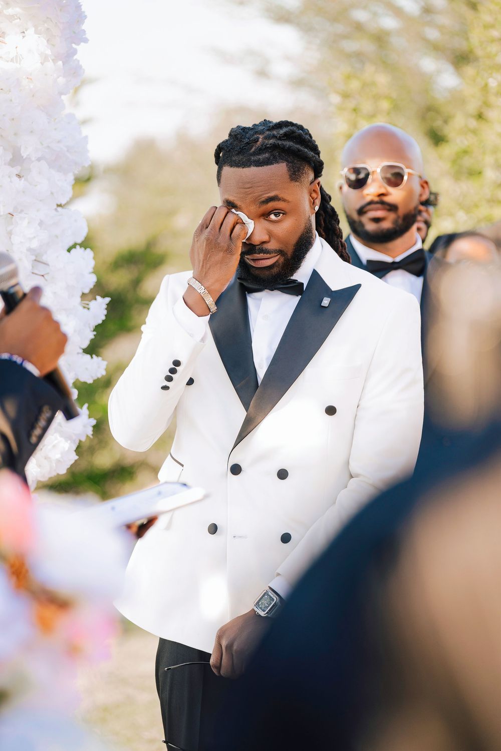 Emotional groom in white tuxedo wiping tears at wedding ceremony with groomsmen standing behind him.