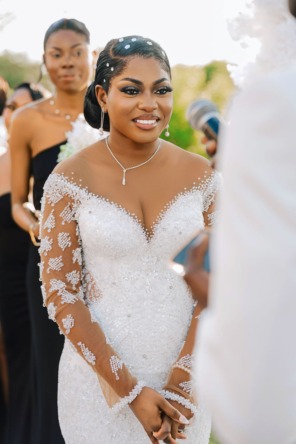 Smiling bride in beaded white gown with sheer sleeves and diamond necklace at outdoor wedding ceremony.
