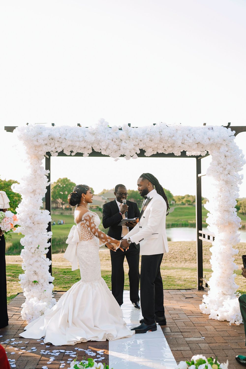 Bride and groom exchange vows under white floral arch at outdoor waterfront wedding ceremony.